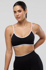 A woman modeling a black sports bra and matching high-waisted leggings with thin white trim, posed against a solid light gray studio background.